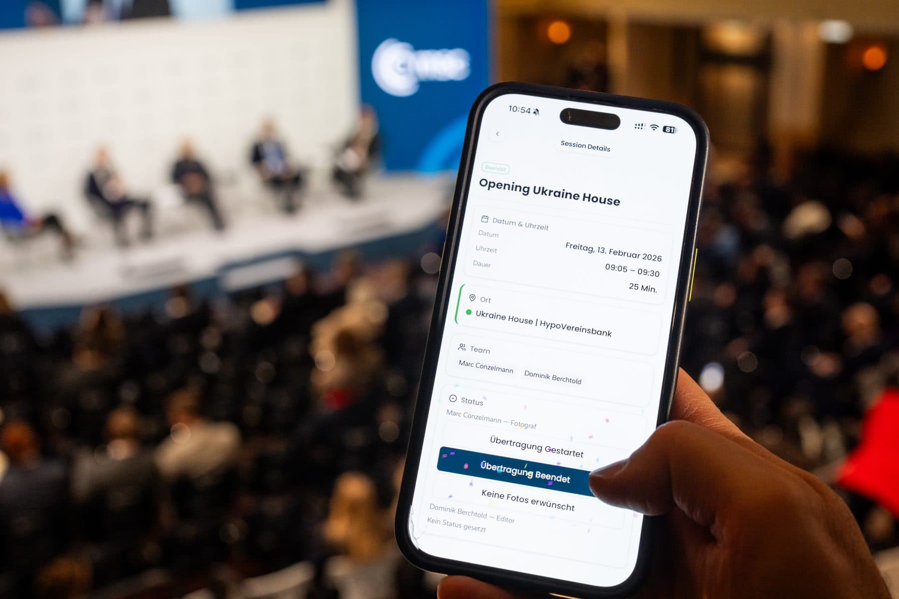 Ein Handy in der Hand im Konferenzsaal: die Session-Detailansicht in VAULO Events mit Termin, Ort, Team und Status — dahinter unscharf das Podium