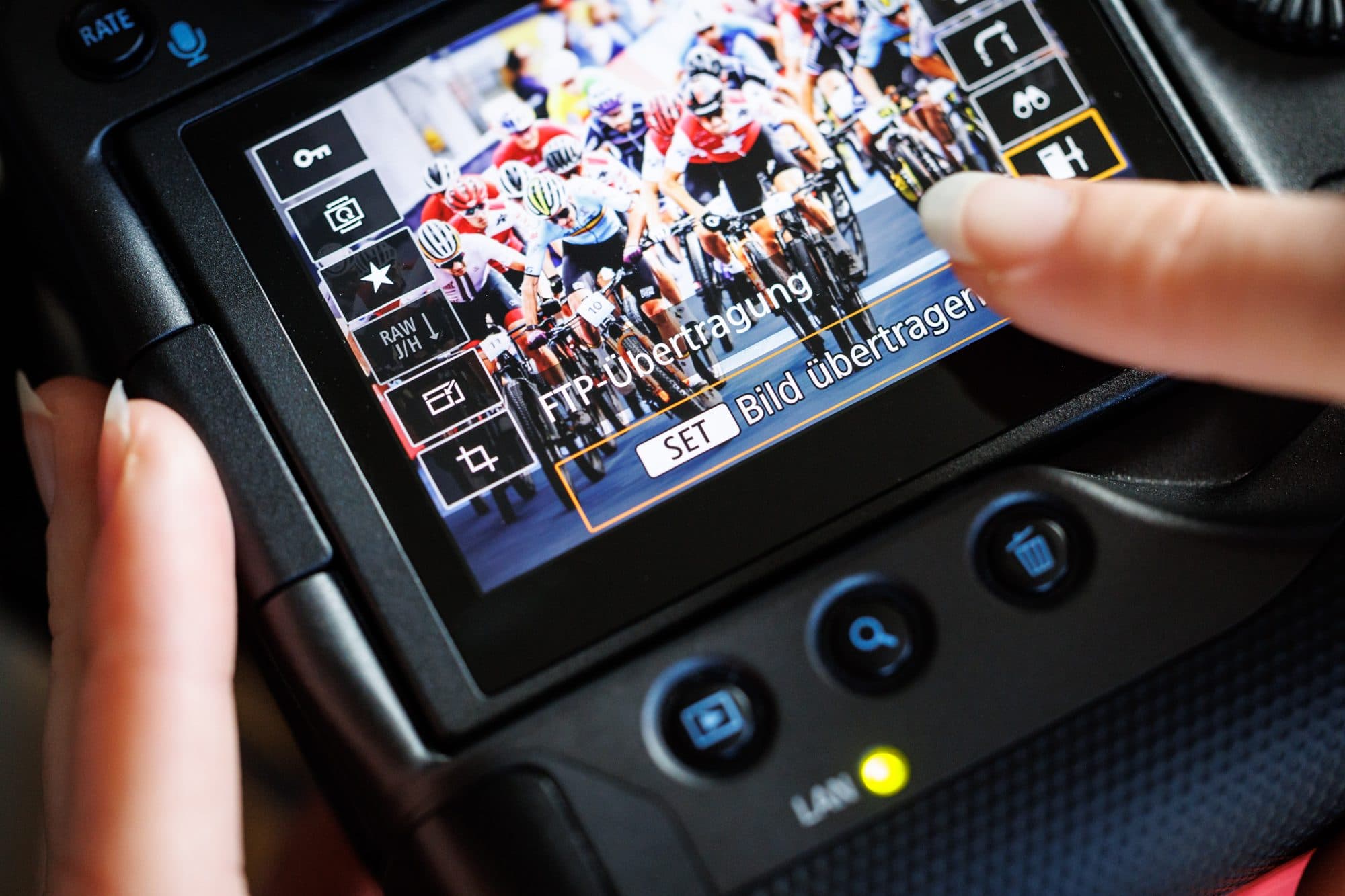 Kamera-Display bei einem Radrennen: das Menü „FTP-Übertragung — Bild übertragen“ liegt über dem Foto des Fahrerfelds, ein Finger tippt auf Übertragen