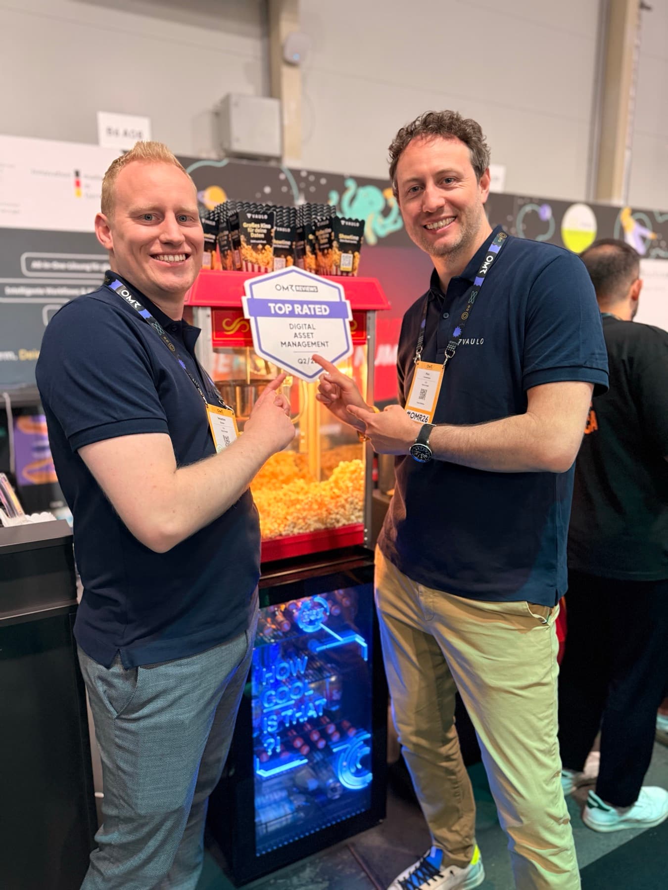 Marc Conzelmann und Sebastian Küsters am VAULO-Messestand — sie zeigen auf das OMR-Reviews-Schild „Top Rated Digital Asset Management“